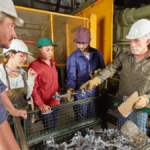 Lean trainer in a heavy-industry plant reviewing cast-metal parts with a diverse production team around a scrap bin.