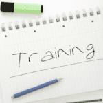 Notebook with the word ‘Training’ handwritten among pens, highlighters, and a drink on a bright desk, representing informal lean on line training preparation.”