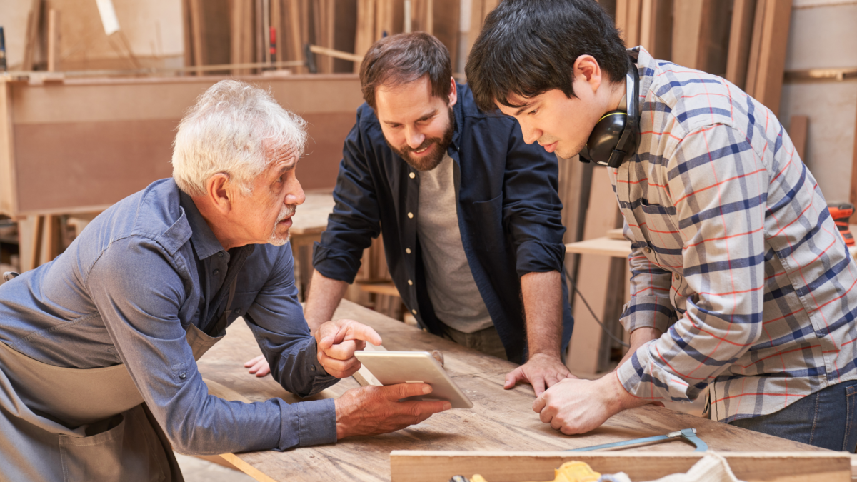 Lean trainer in a small woodworking shop guiding two younger colleagues with a digital tablet at a wooden workbench.