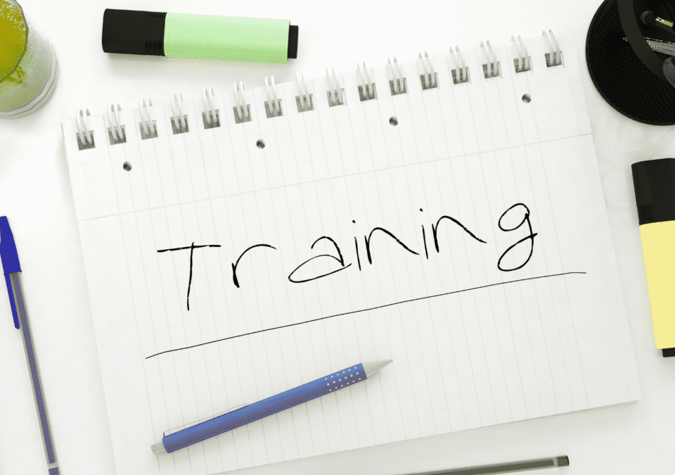 Notebook with the word ‘Training’ handwritten among pens, highlighters, and a drink on a bright desk, representing informal lean on line training preparation.”